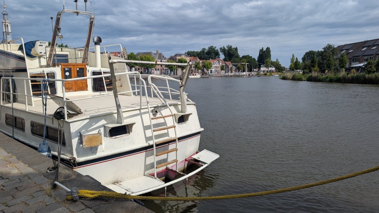 Witte boot aangemeerd langs een kanaal met huizen en bomen op de achtergrond onder een bewolkte hemel.