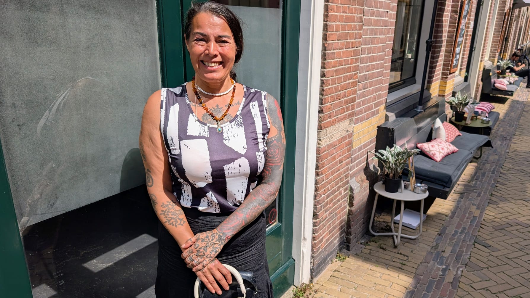 Vrouw glimlacht voor een groen deurkozijn, terwijl ze een handtas vasthoudt; straat met zitjes op de achtergrond.
