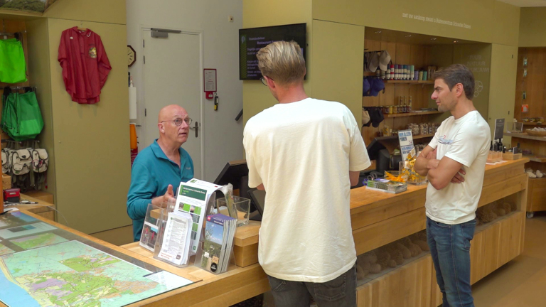 Drie mannen staan bij een balie in een winkel met kaarten en wandelsouvenirs op de achtergrond.