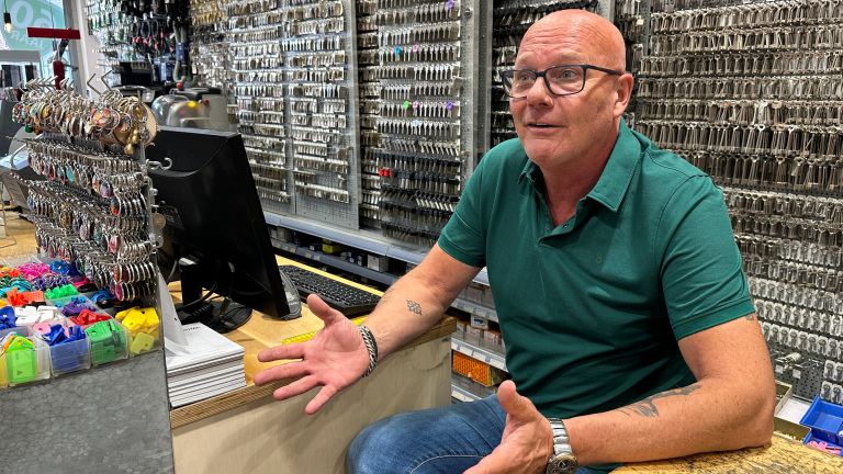 Man bij een balie in een winkel vol sleutels, pratend en gebarend, omringd door diverse sleutelhangers en computer.