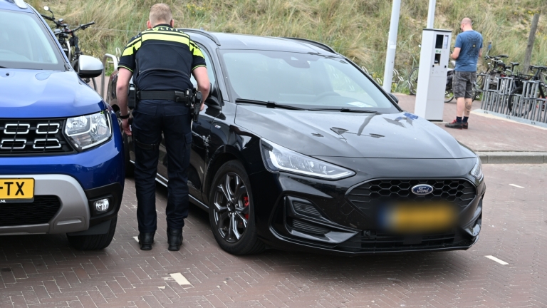 Een politieagent controleert een zwarte auto op een parkeerplaats, met een felblauwe auto naast zich.