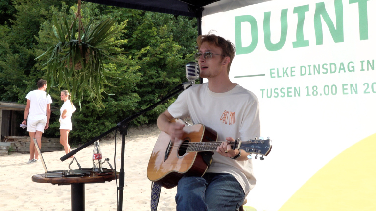 Man speelt gitaar en zingt bij een microfoon onder een tent, met achtergrond van bomen; tekst vermeldt een evenementijd.