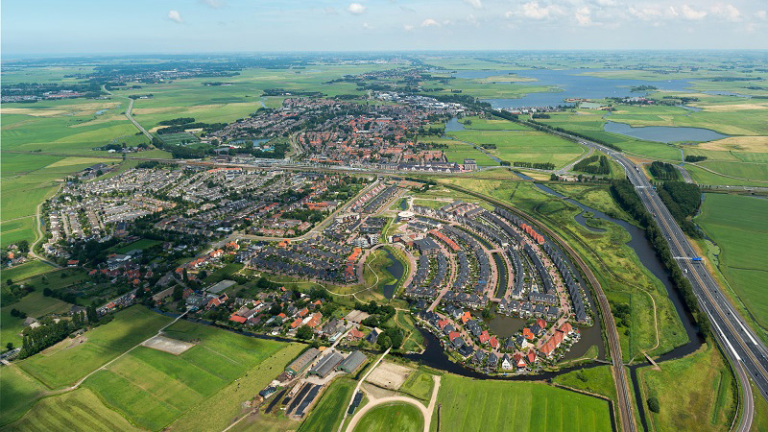 Luchtfoto van een uitgestrekt groen landschap met een woonwijk. Slingerweg en waterwegen zijn zichtbaar tussen de huizen.