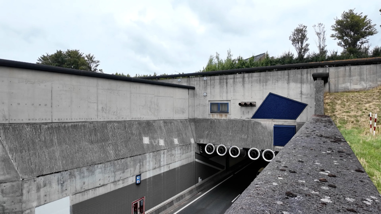 Ingang van een verkeerstunnel met betonnen wanden, blauwe ventilatieroosters en afwateringsbuizen onder een bewolkte lucht