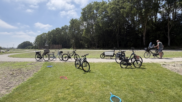 Fietsen staan langs een pad in een park met bomen; rechts duwt een man een fiets met kinderzitje