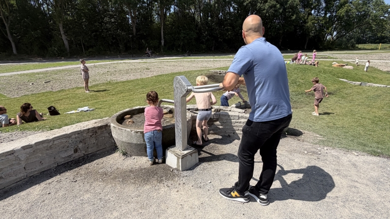 Man helpt kinderen bij een waterpomp naast een stenen bak in een park, met spelende mensen op het gras