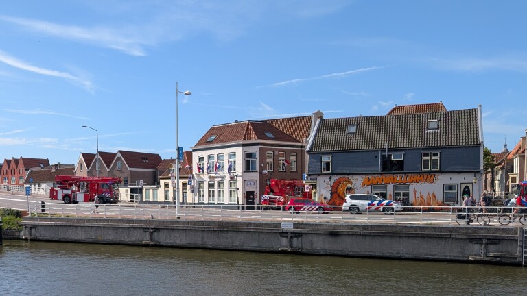 Brandweerwagens en politiewagen op brug bij kanaal, met gevelmuur “AANVALLUHH!!” en fietsers op de weg