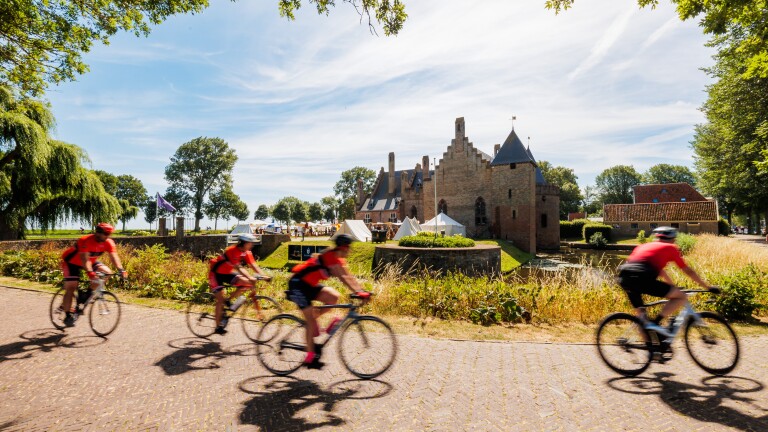 Wielrenners in rode shirts rijden langs een middeleeuws kasteel met gracht en tenten op een zonnige dag