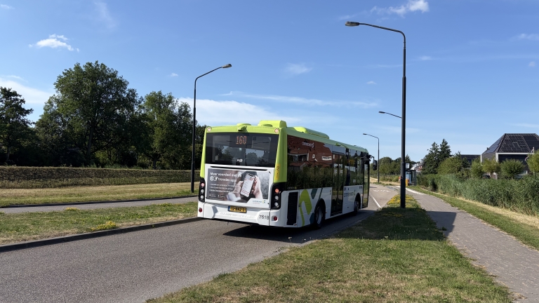 Groen-witte lijnbus rijdt over een rustige weg langs gras en bomen, met halte en straatlantaarns onder blauwe lucht