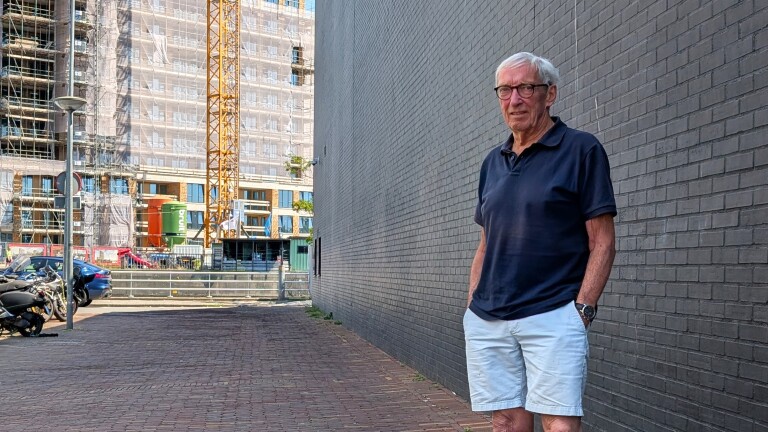 Oudere man met bril leunt tegen een bakstenen muur, met bouwplaats en hijskraan op de achtergrond