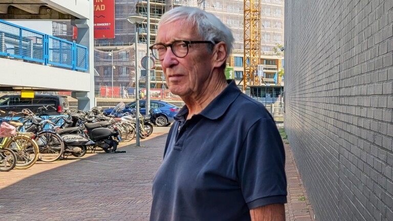 Oudere man met grijs haar en bril in donkerblauwe polo, staand langs een bakstenen muur bij fietsen en bouwplaats
