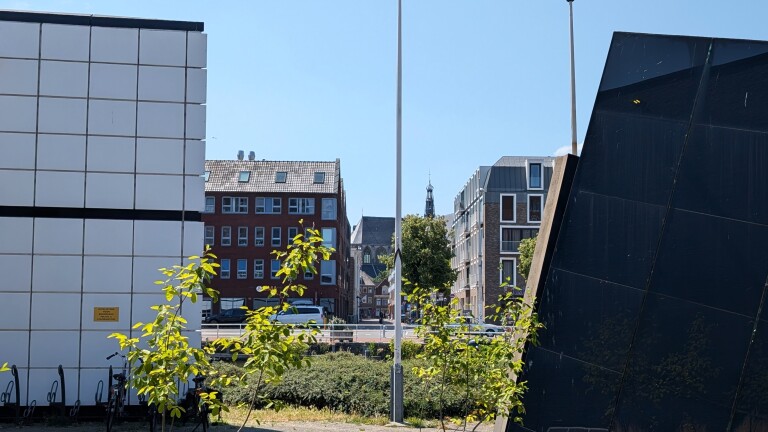 Stadszicht tussen twee moderne gebouwen, met jonge boompjes, geparkeerde fietsen en huizen met kerktoren op de achtergrond