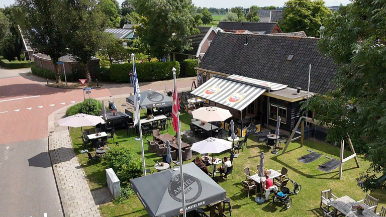 Terras met tafels en parasols naast een café met gestreepte luifel, aan een kruispunt met veel groen
