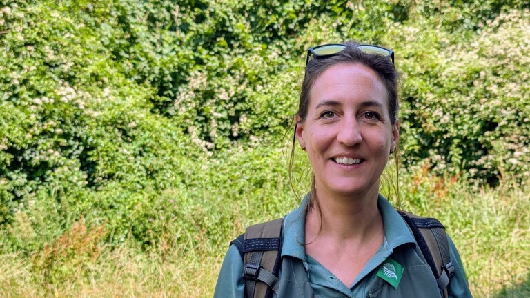 Vrouw met rugzak en zonnebril op het hoofd glimlacht voor een groene, bosrijke achtergrond