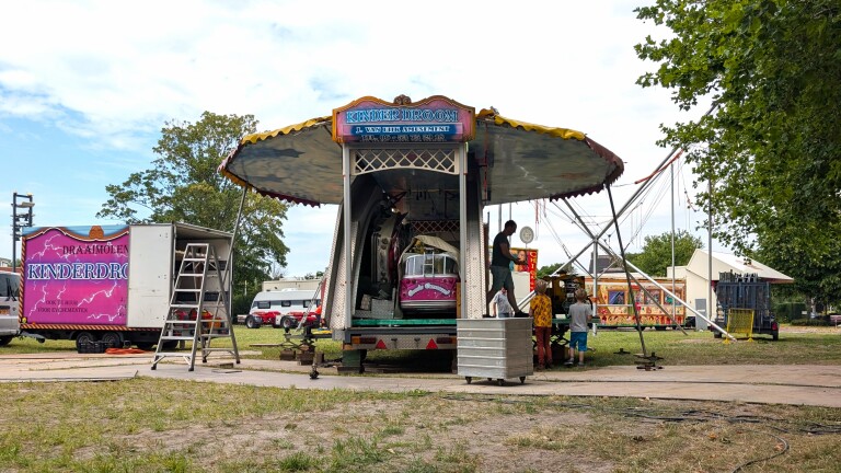 Opbouw van kermisattractie KinderDroom met open trailer, ladder en kinderen bij een medewerker op het terrein