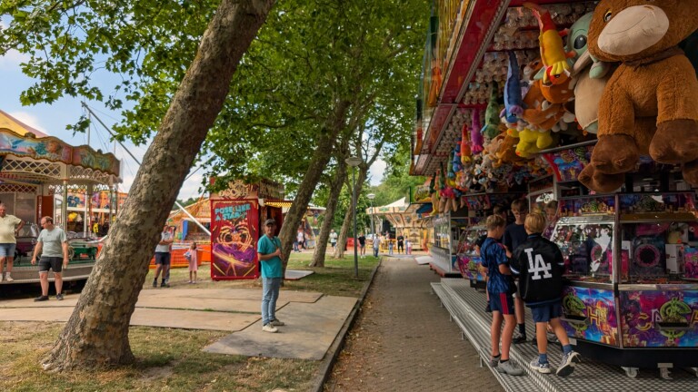 Kermis onder bomen, met bezoekers bij een grijpkraam vol knuffels en attracties op de achtergrond