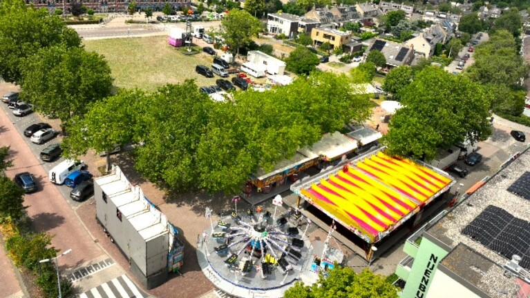 Luchtfoto van een kermis met draaimolen en geel-rood gestreepte tent tussen bomen, naast straten en huizen