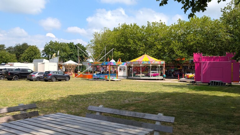 Kermisattracties op een grasveld met draaimolen, kraampjes en geparkeerde auto’s, met bomen en blauwe lucht