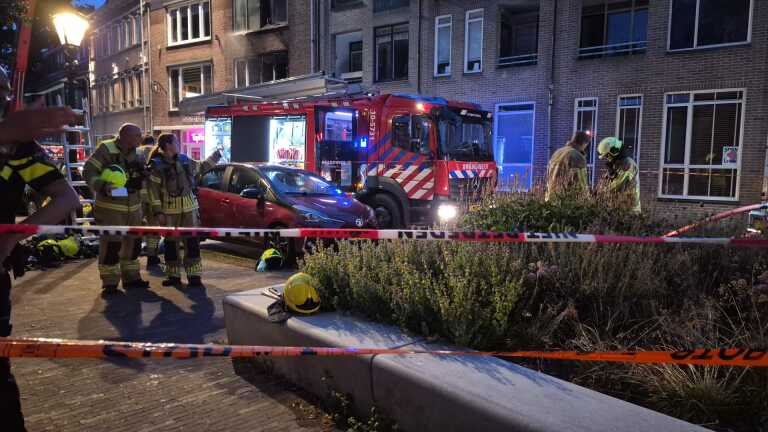 Brandweerwagens en brandweerlieden achter afzetlint bij een gebouw met rookschade en kapotte ramen in de avond