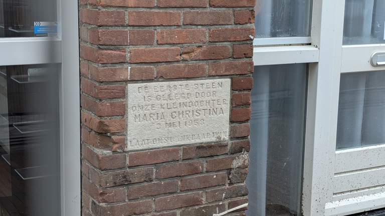 Gedenksteen in bakstenen muur bij een deur, met gegraveerde tekst over de eerste steen gelegd op 3 mei 1958