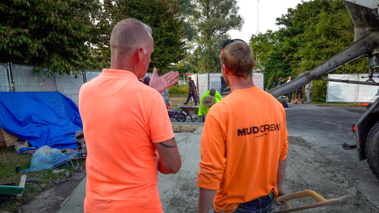 Twee bouwvakkers in oranje shirts (MUD CREW) bekijken betonstort terwijl een betonpomp beton uitgiet