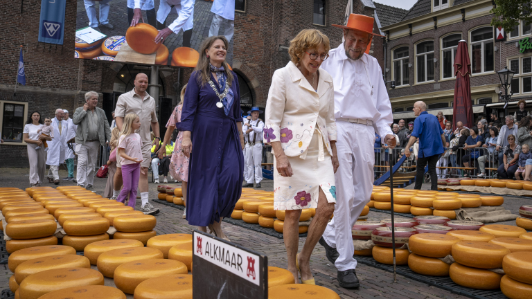 Mensen lopen over rijen ronde kazen op een plein in Alkmaar, met publiek en een groot scherm op de achtergrond