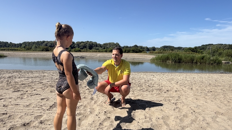 Badmeester in geel shirt knielt op het strand en geeft een handdoek aan een kind bij een rustig meer