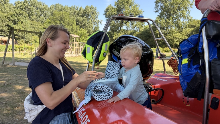 Vrouw geeft peuter een voorwerp terwijl hij op een rode reddingsboot zit, met reddingsvesten en bomen op de achtergrond