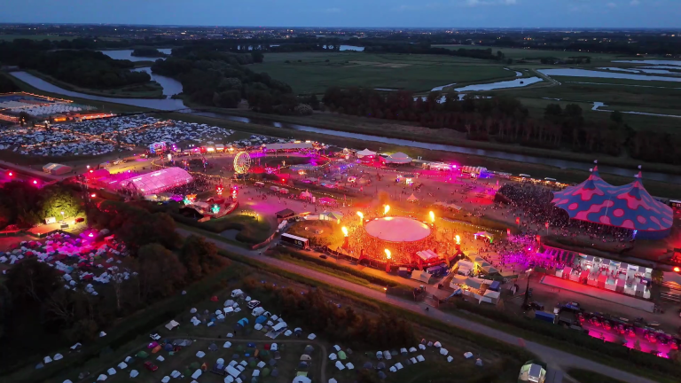 Luchtfoto van festivalterrein in de schemering met felgekleurde lichten, groot tentdak, reuzenrad en camping eromheen