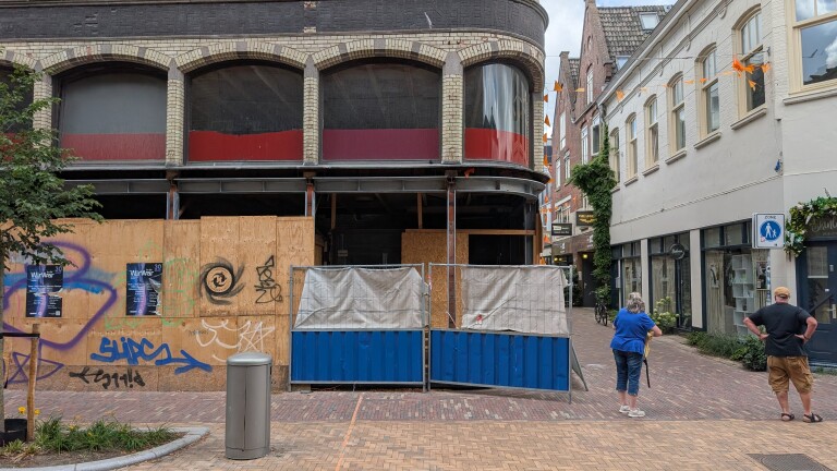 Straat met verbouwing: pand met houten schotten en graffiti, afzethekken, en twee voorbijgangers op de stoep