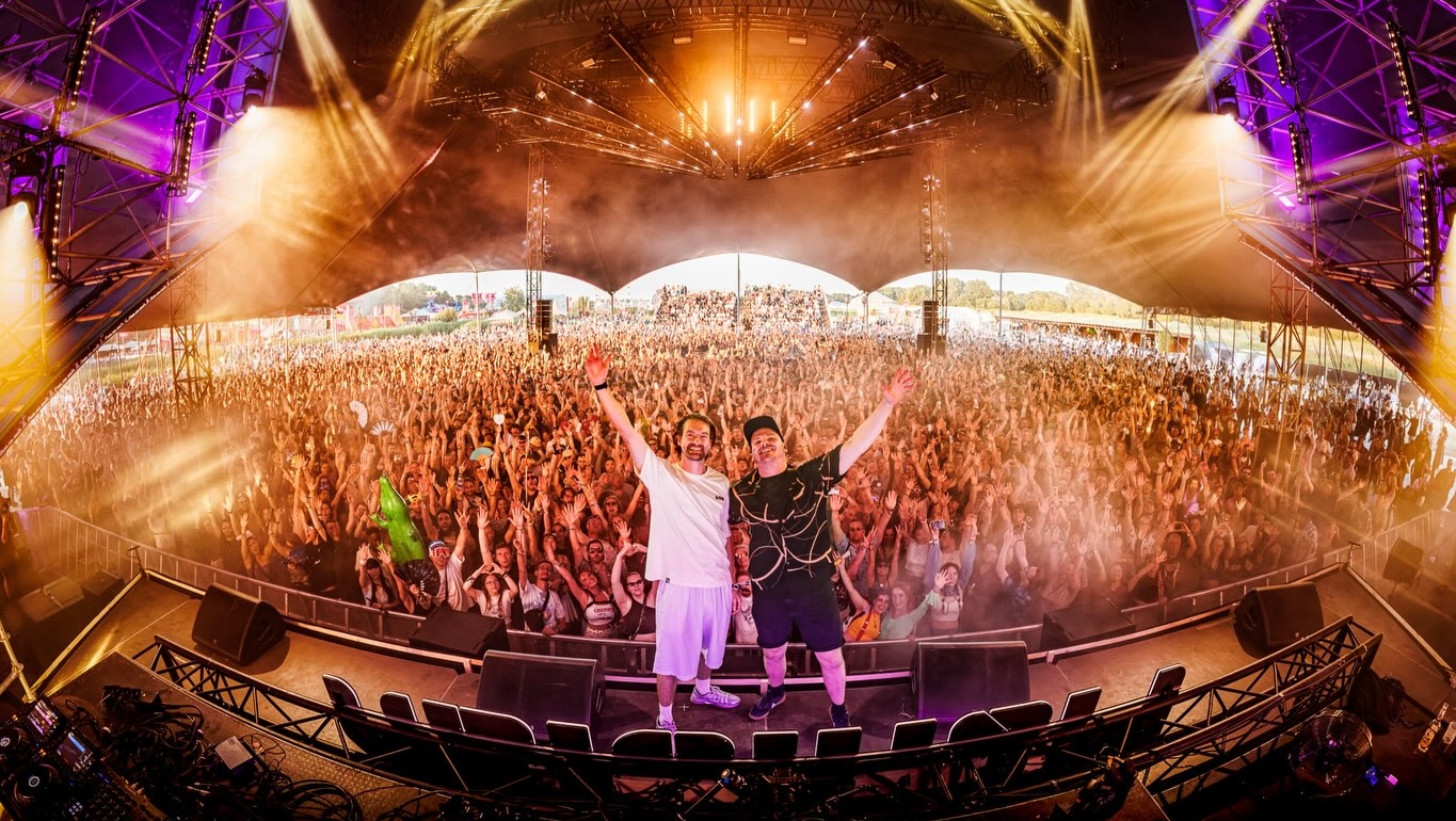 Twee dj’s op een festivalpodium met armen omhoog, voor een enorme juichende menigte onder paarse en gouden lichten