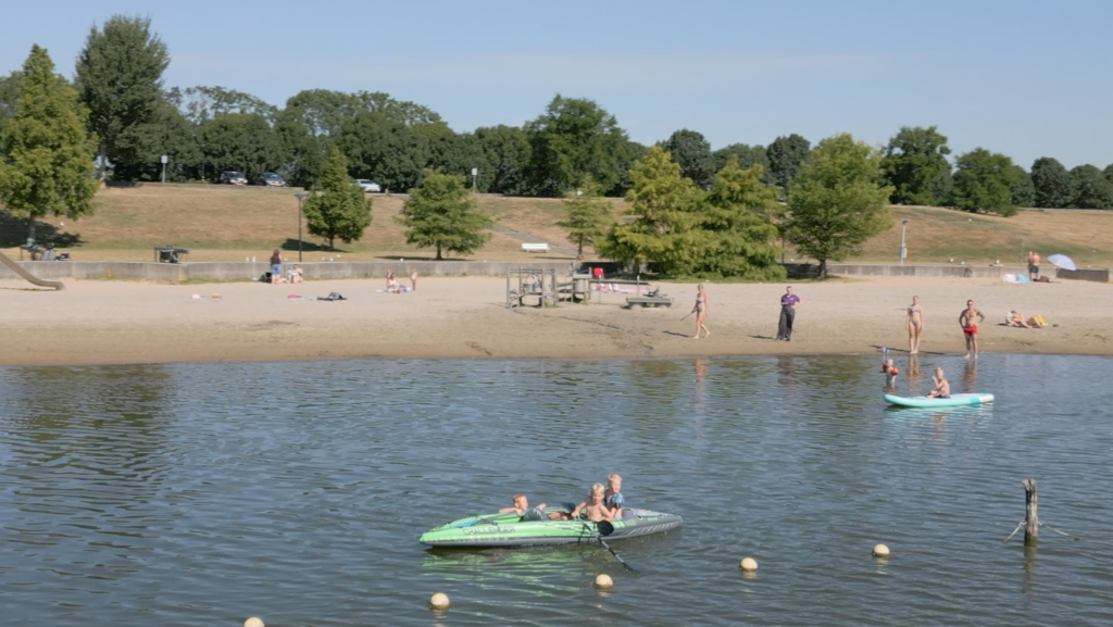 Kinderen in een opblaasboot op een meer, met een zandstrand en badgasten op de achtergrond