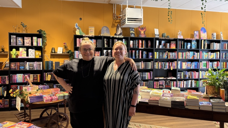 Twee vrouwen poseren in een boekwinkel voor volle boekenkasten, met stapels boeken op een tafel op de voorgrond