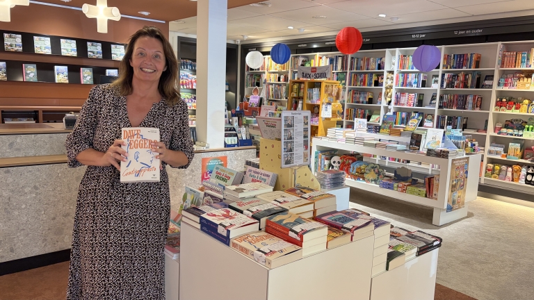 Vrouw in boekwinkel houdt het boek Contrapasso van Dave Eggers vast, met boekentafels en kleurrijke lampionnen op de achtergrond