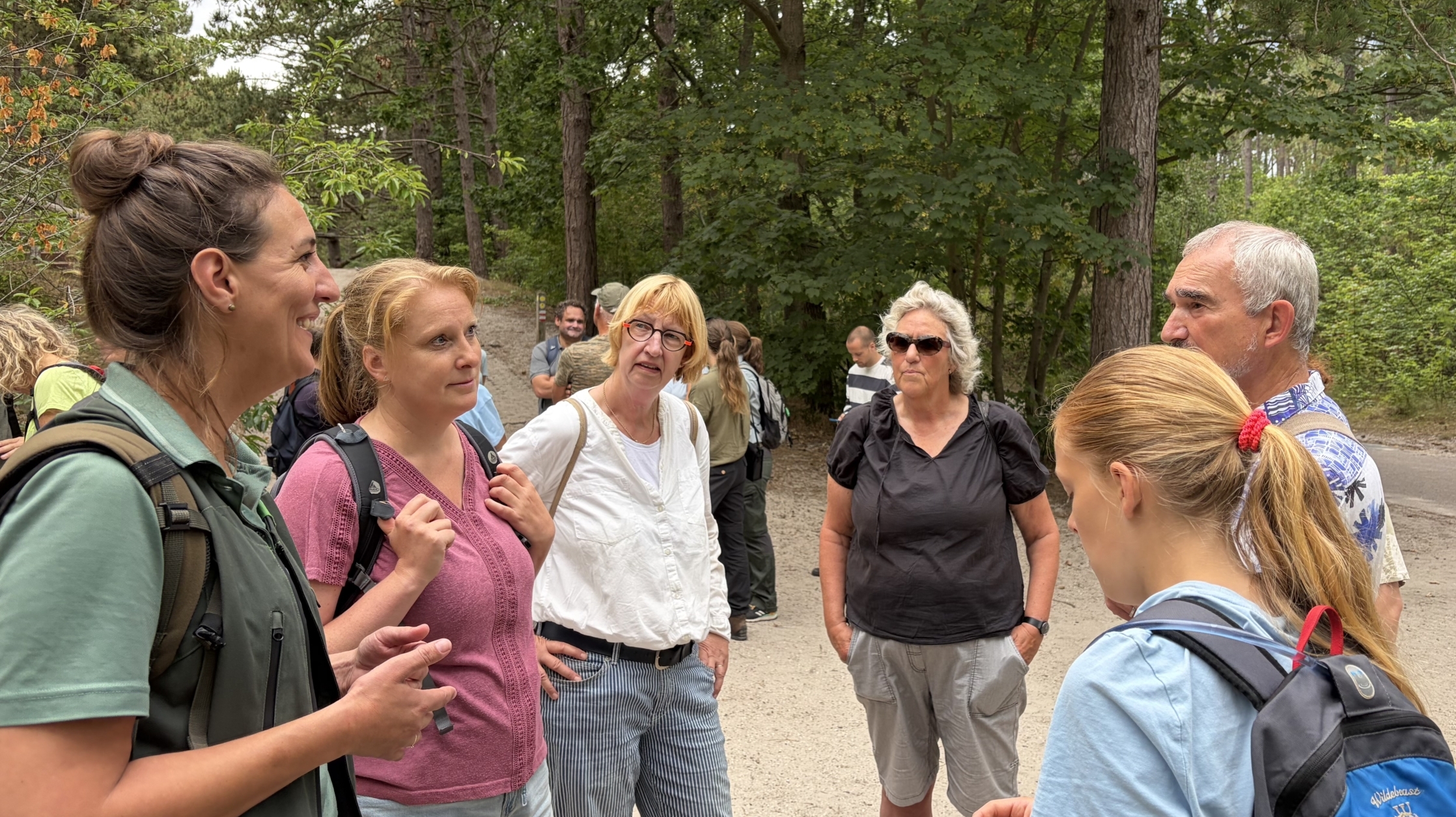 Groep wandelaars met rugzakken staat te praten bij een bospad, omringd door hoge bomen