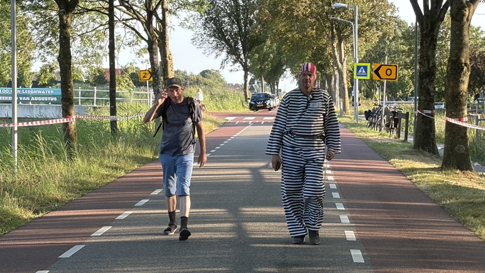 Twee mannen lopen op een fietspad langs bomen; één draagt een zwart-wit gestreept gevangenispak