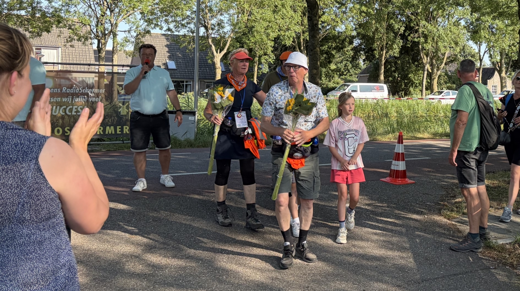 Wandelaars met bloemen worden toegejuicht bij een evenement op een boomrijke weg, met ballonnen en bord “BIJ ZUID”