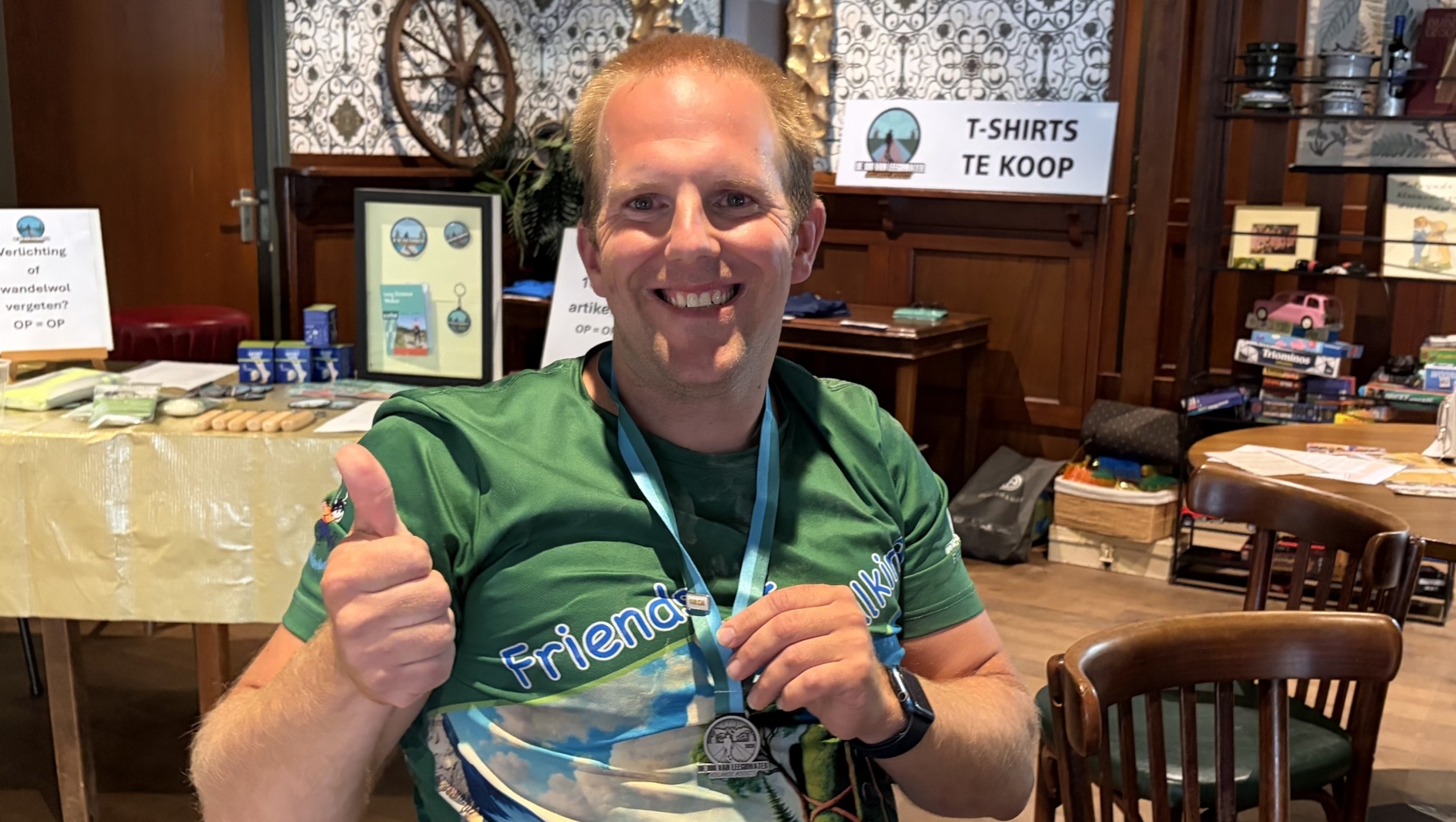 Glunderende man in groen shirt met medaille geeft duim omhoog aan tafel met verkoopstand op de achtergrond