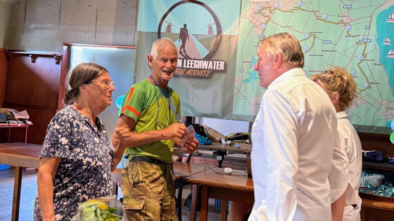 Drie mensen praten in een zaal voor een kaart en banner van “Van Leeghwater Lands Mooiste”