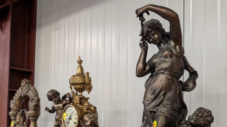 Bronzen beeld van een vrouw met opgeheven arm, naast een sierlijke gouden mantelklok met cherubijnen