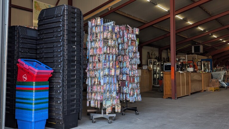 Stapel zwarte en gekleurde winkelmanden naast een rek vol sleutelhangers in een loods met tweedehands spullen