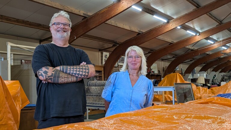 Twee glimlachende mensen in een loods tussen met oranje zeilen afgedekte meubels, met houten spanten boven hen