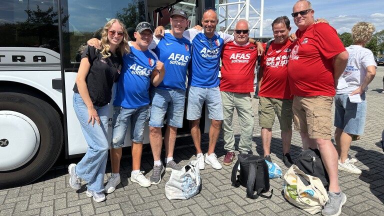 Groep supporters in blauwe en rode AZ-shirts poseert arm in arm naast een bus op een zonnige dag