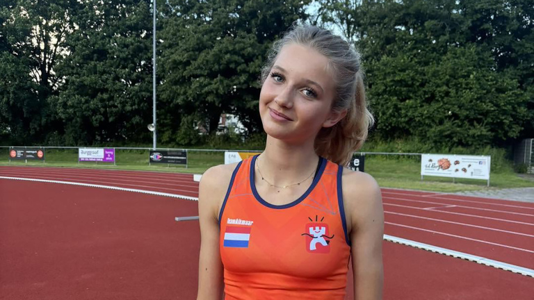 Atlete in oranje top met Nederlandse vlag staat op atletiekbaan, met bomen en reclameborden op de achtergrond