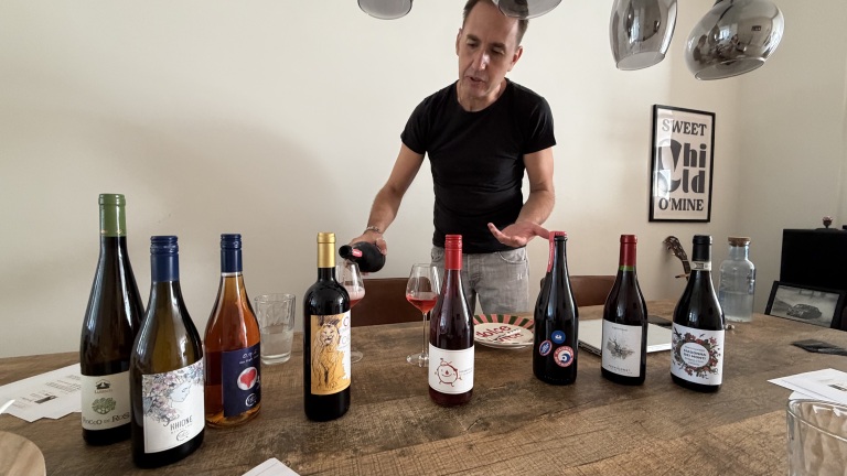 Man schenkt rosé in wijnglas aan tafel met meerdere wijnflessen tijdens een proeverij in een woonkamer