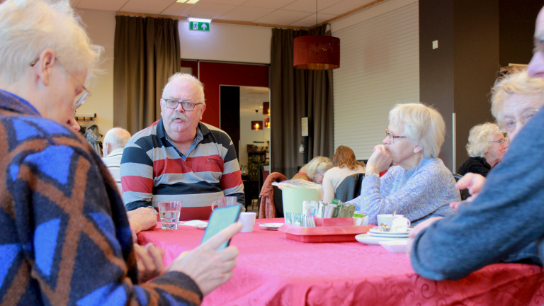 Groep ouderen zit aan een tafel met rood tafelkleed en koffiekopjes in een ontmoetingsruimte, in gesprek