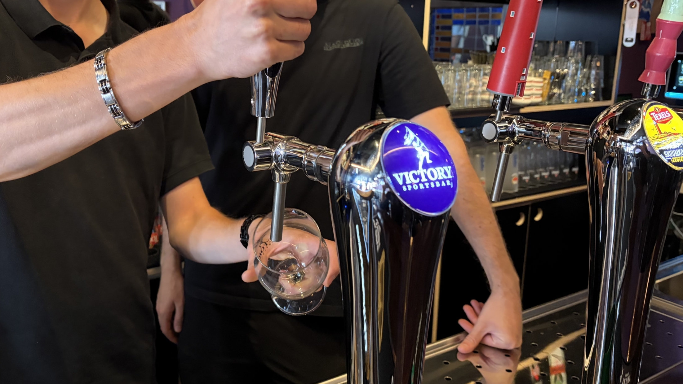 Barman tapt bier in een glas aan de bar, met glanzende tapkranen en het logo “Victory Sportsbar” zichtbaar