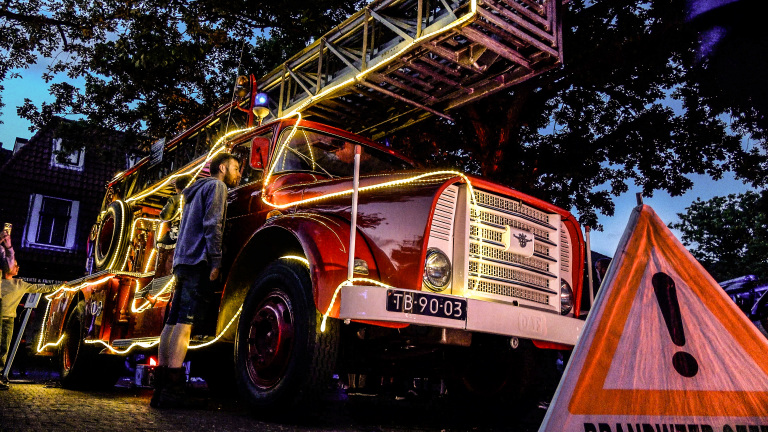 Rode brandweerwagen met uitgeschoven ladder, versierd met lichtsnoeren, terwijl mensen er ’s avonds naast staan