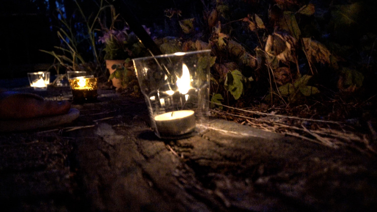 Brandend waxinelichtje in een glazen windlicht op een stenen pad, met nog twee kaarsjes en planten op de achtergrond