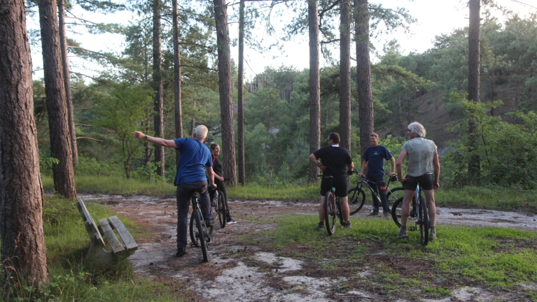 Groep fietsers pauzeert op een bospad tussen hoge dennen, terwijl één man met gestrekte arm de route aanwijst